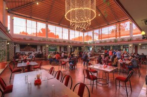 A spacious indoor dining area with multiple tables and chairs, large windows, and a decorative chandelier. - GRACIAN Heritage Shelters Resort  (Shrirangapattana)