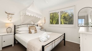 A bedroom with a canopy bed, a bedside table with a lamp, a window with blinds, and a mirror on the wall. - The Address by Experience Jervis Bay (Vincentia)