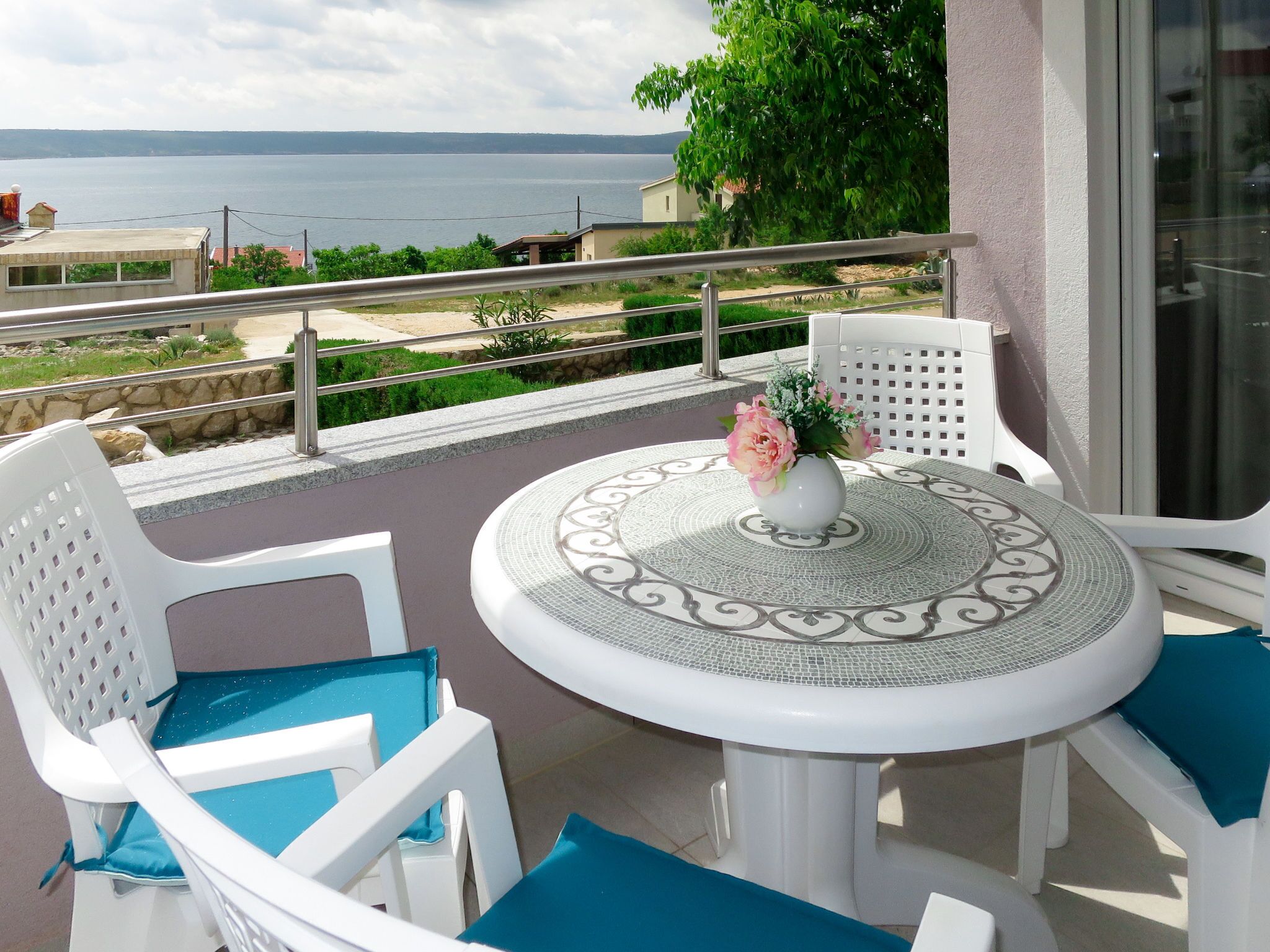 Una mesa redonda con un jarrón blanco lleno de flores, rodeada de sillas blancas con cojines azules, en un patio exterior con vistas al mar.