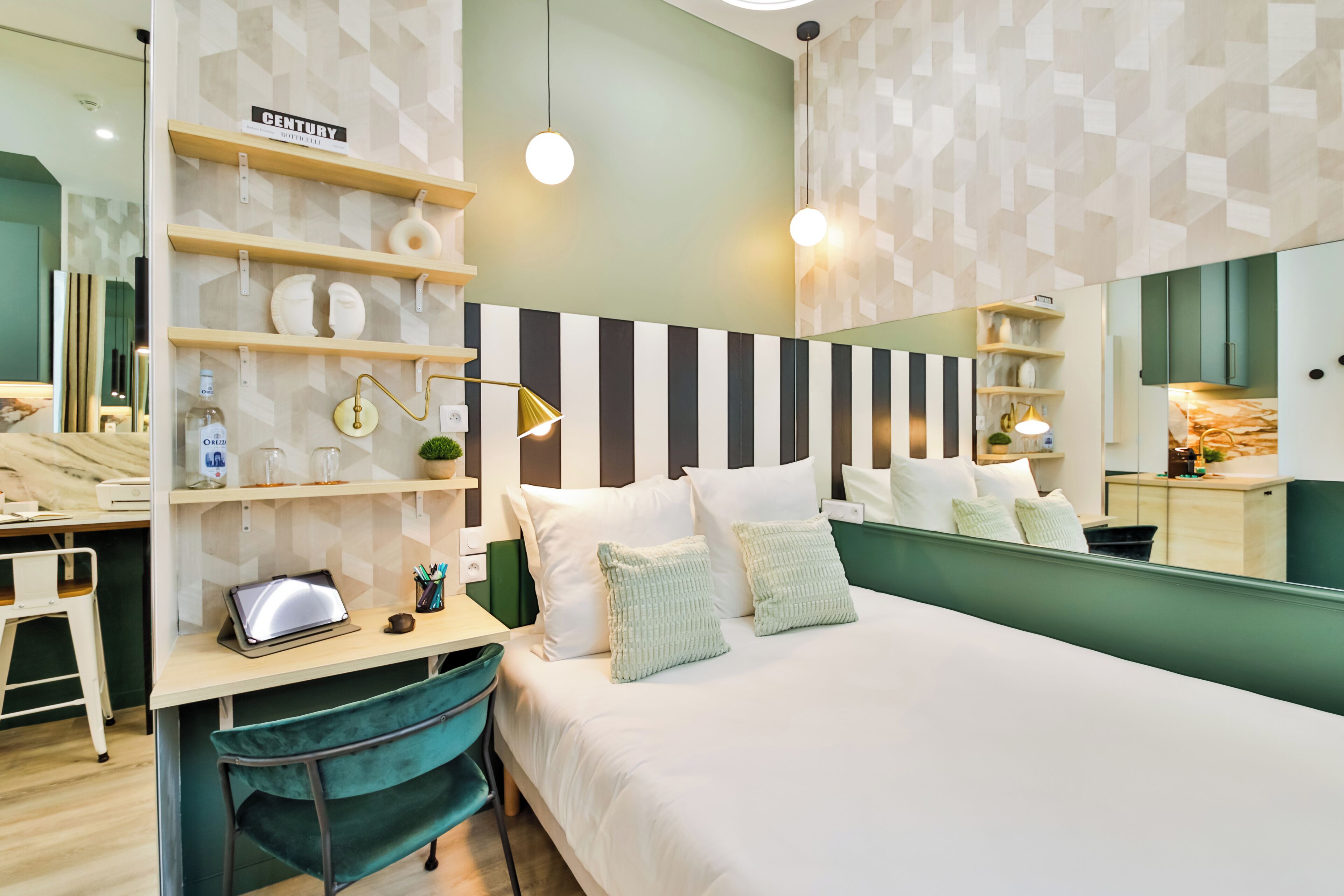 A modern hotel room with a bed, a desk with a laptop, shelves with decorative items, and a large mirror.