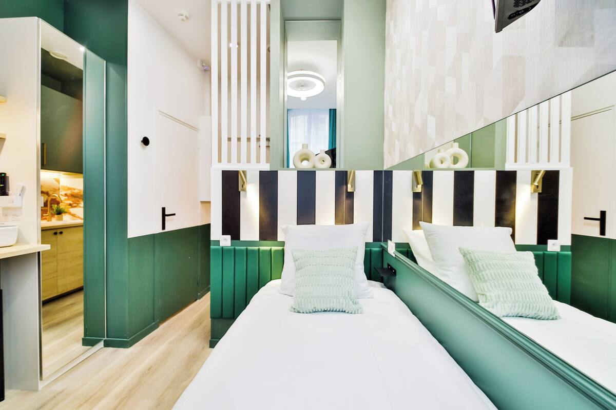 A modern bedroom with a large mirror, two beds, a green accent wall, and a visible kitchenette area.