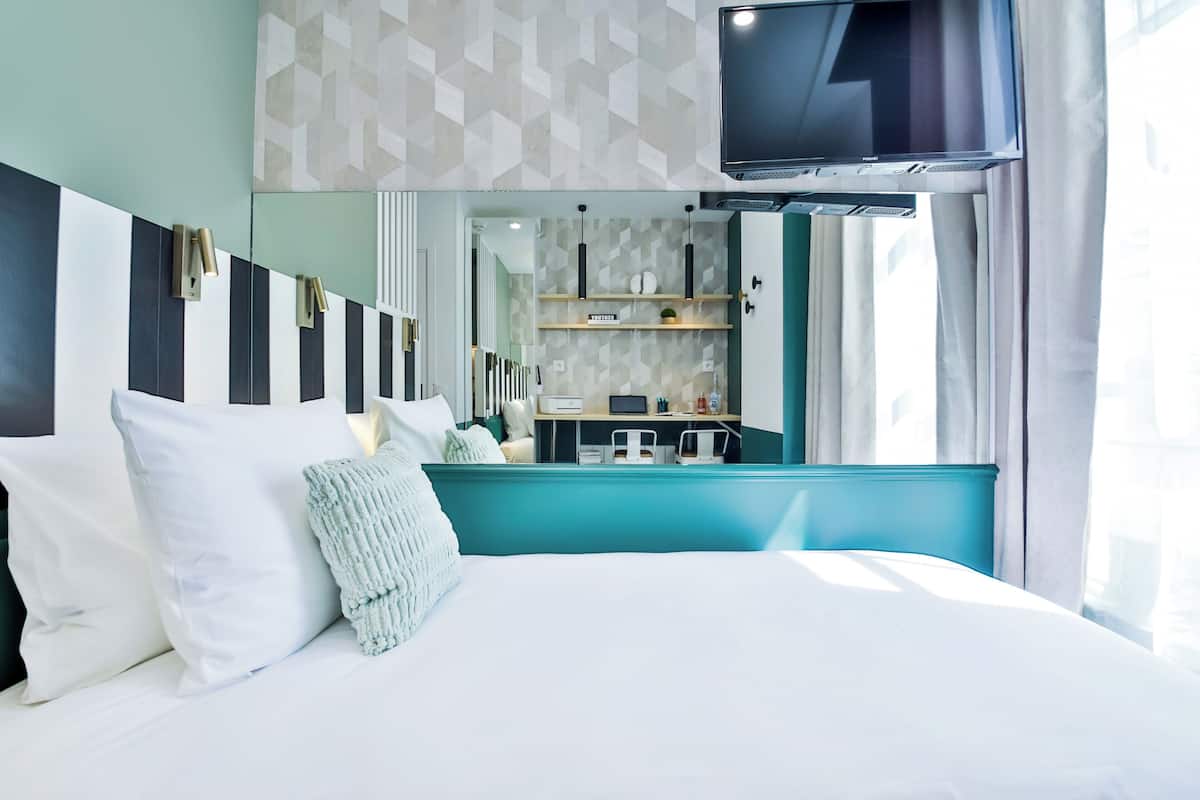 A modern hotel room with a bed, a TV, a mirror, and a kitchenette.