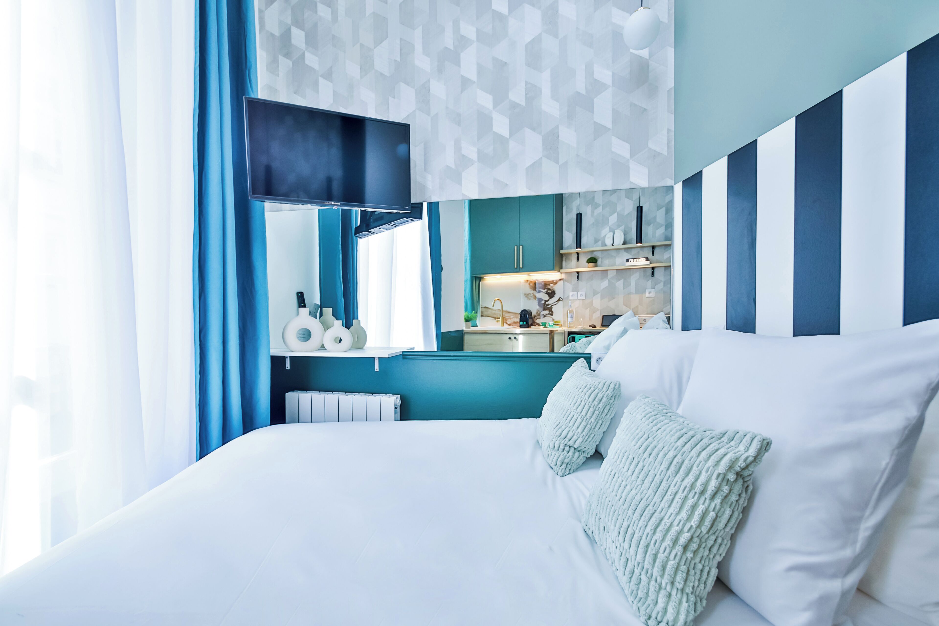 A modern hotel room with a large bed, a view of the kitchen, and a geometric patterned wall.