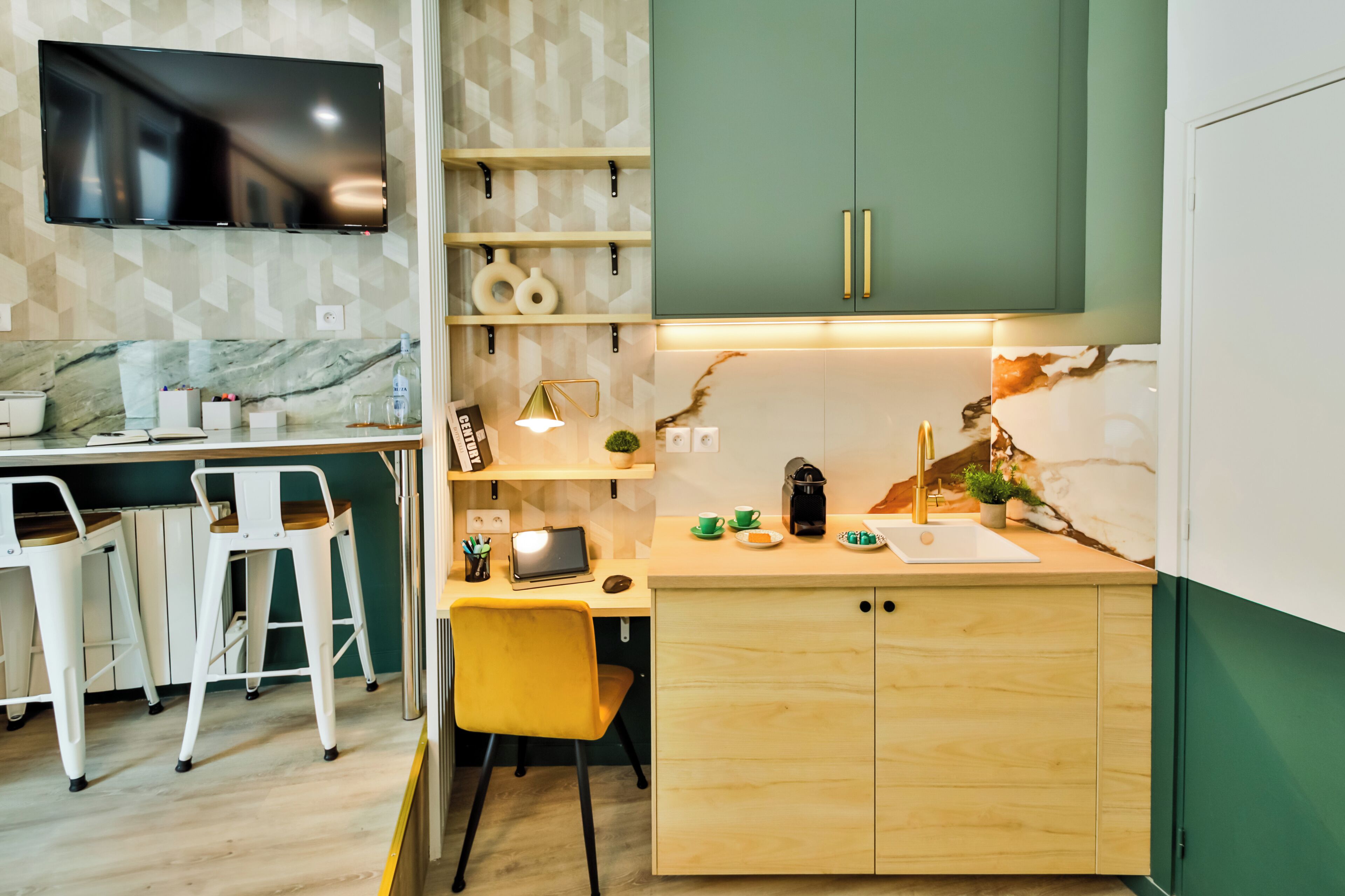 Une cuisine moderne avec un plan de travail en bois, un four encastré et un meuble vert. On y trouve également un coin bar avec des tabourets blancs et un téléviseur fixé au mur.