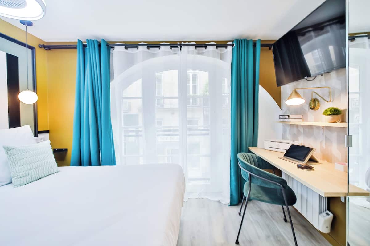 A modern hotel room with a bed, a desk with a laptop, a TV, and a window with curtains.