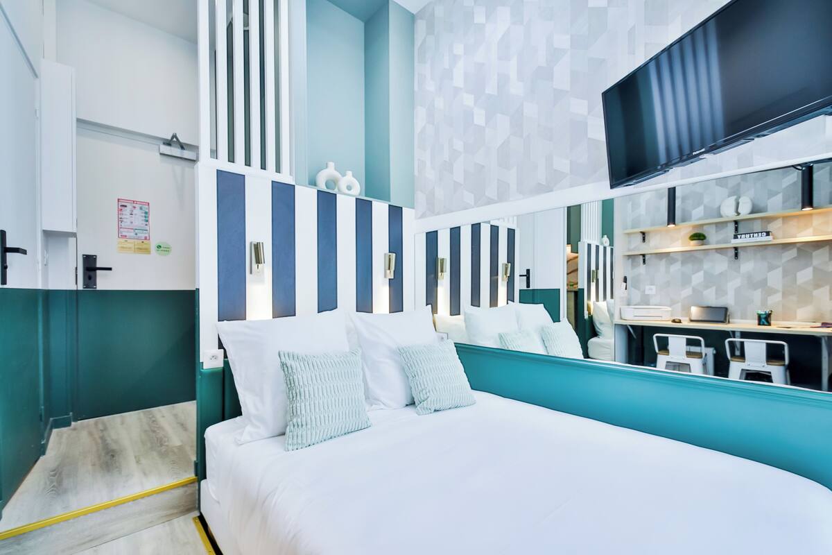 A modern hotel room with a large bed, a TV mounted on the wall, and a small kitchenette area.