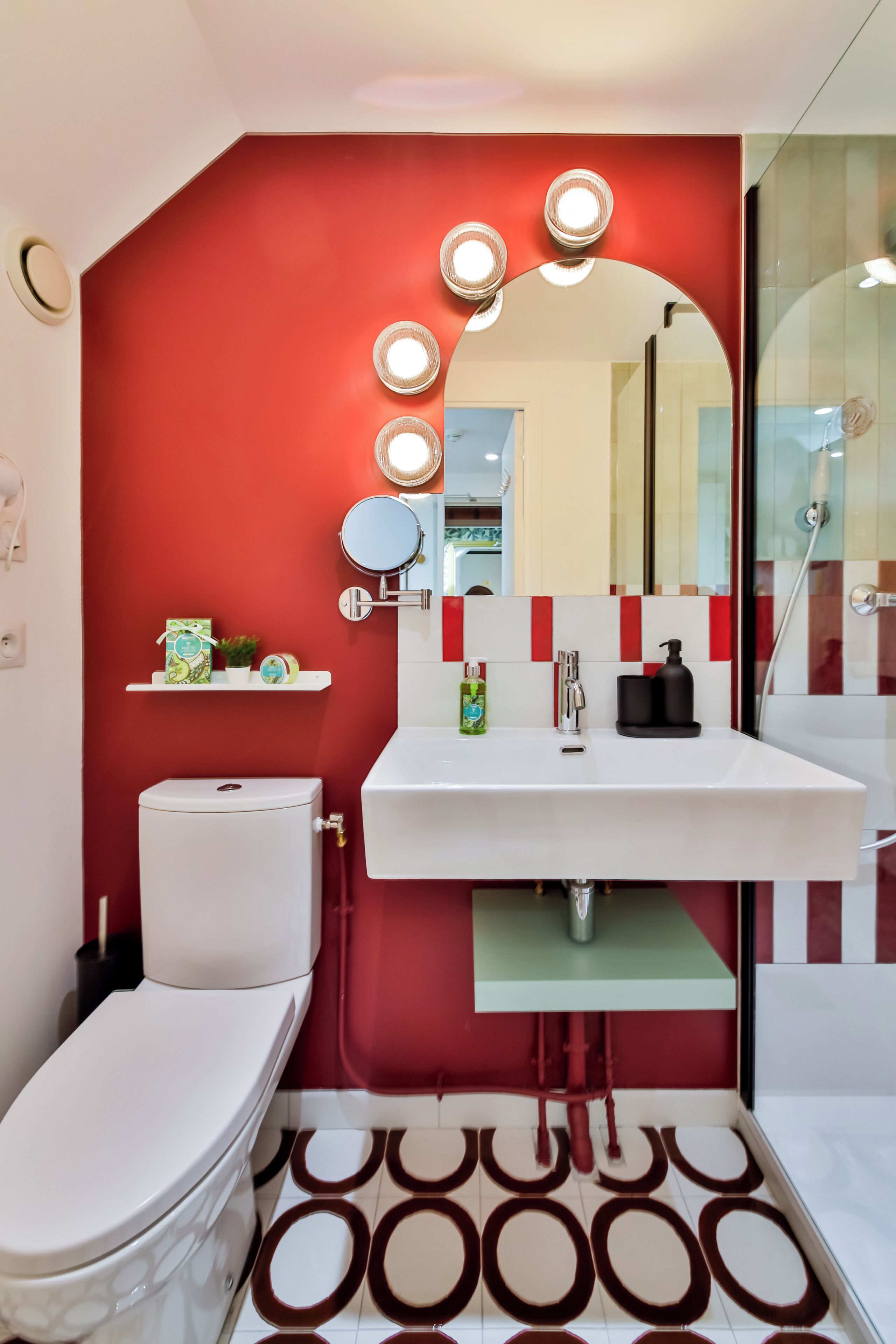 A bathroom with a red accent wall, a white sink, a mirror with lights, a toilet, and patterned floor tiles.