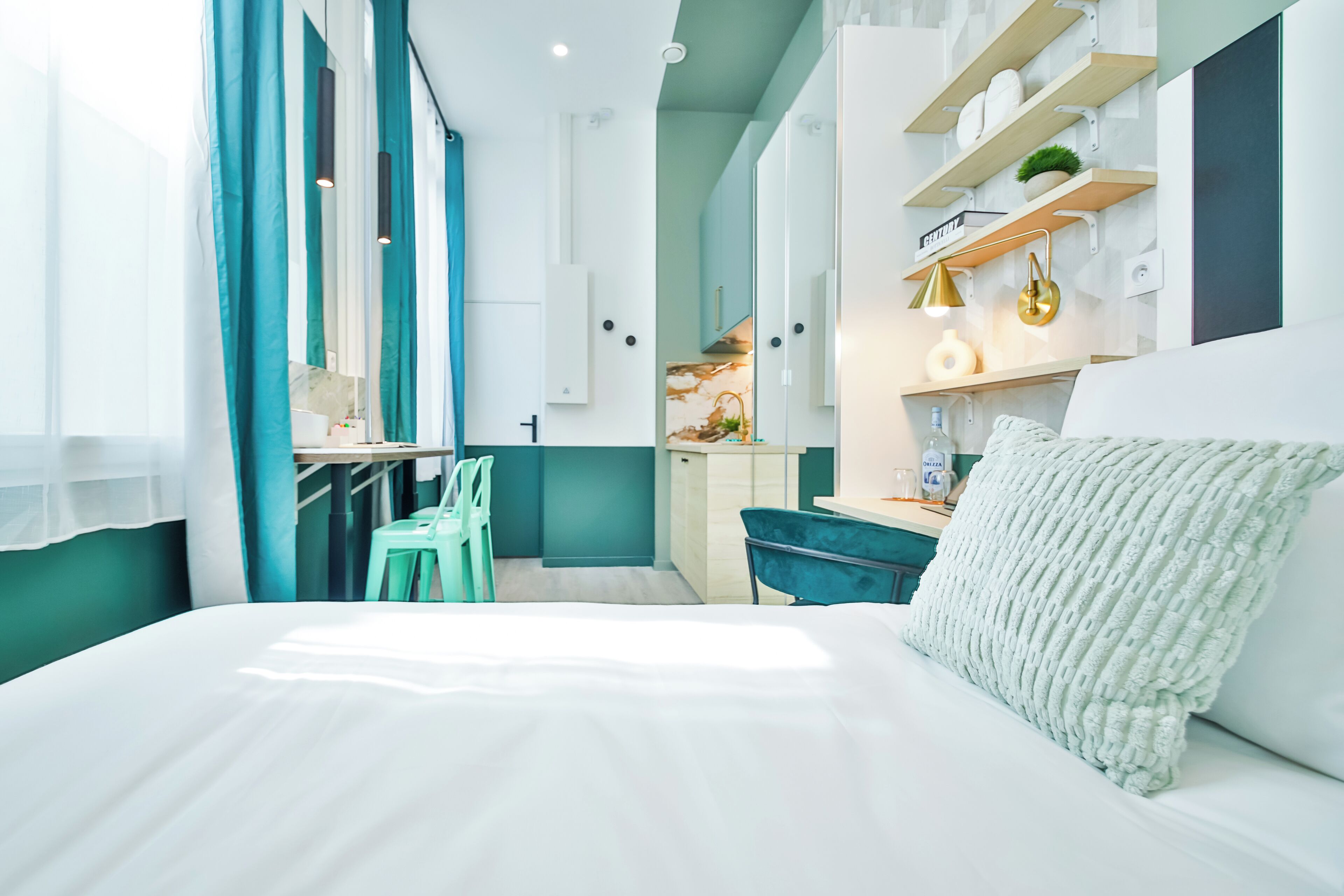 Moderni hotellihuone, jossa on sänky, työpöytä, tuoli ja pieni keittiönurkkaus.