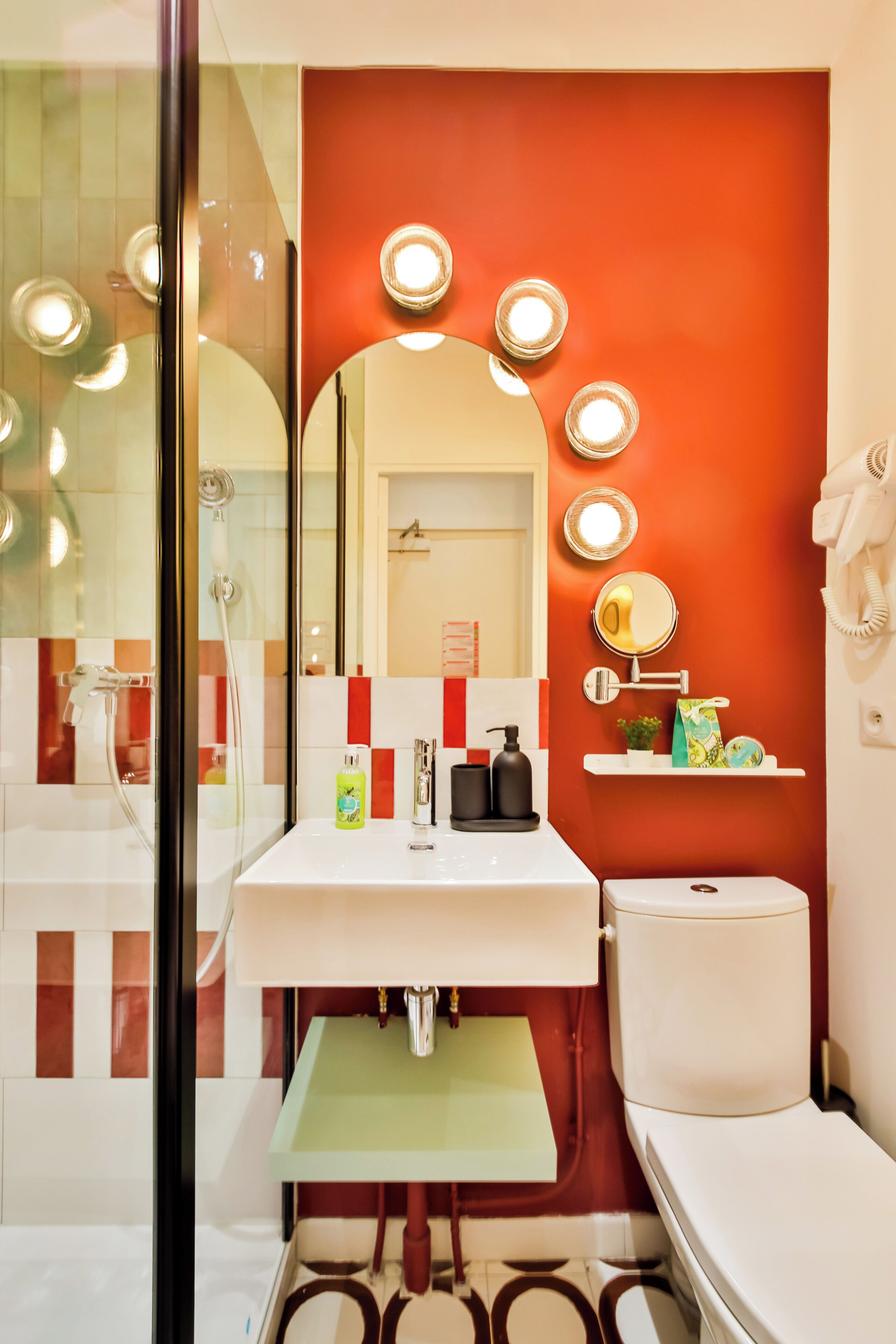 A bathroom with a glass shower enclosure, a white sink with a green vanity, a white toilet, and a red accent wall with a mirror and lights.