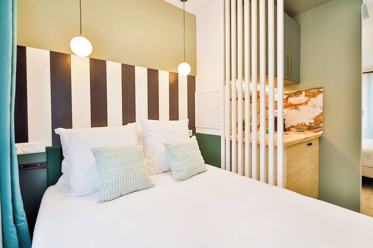 A modern hotel room with a large bed, striped headboard, and a view of the kitchen area.