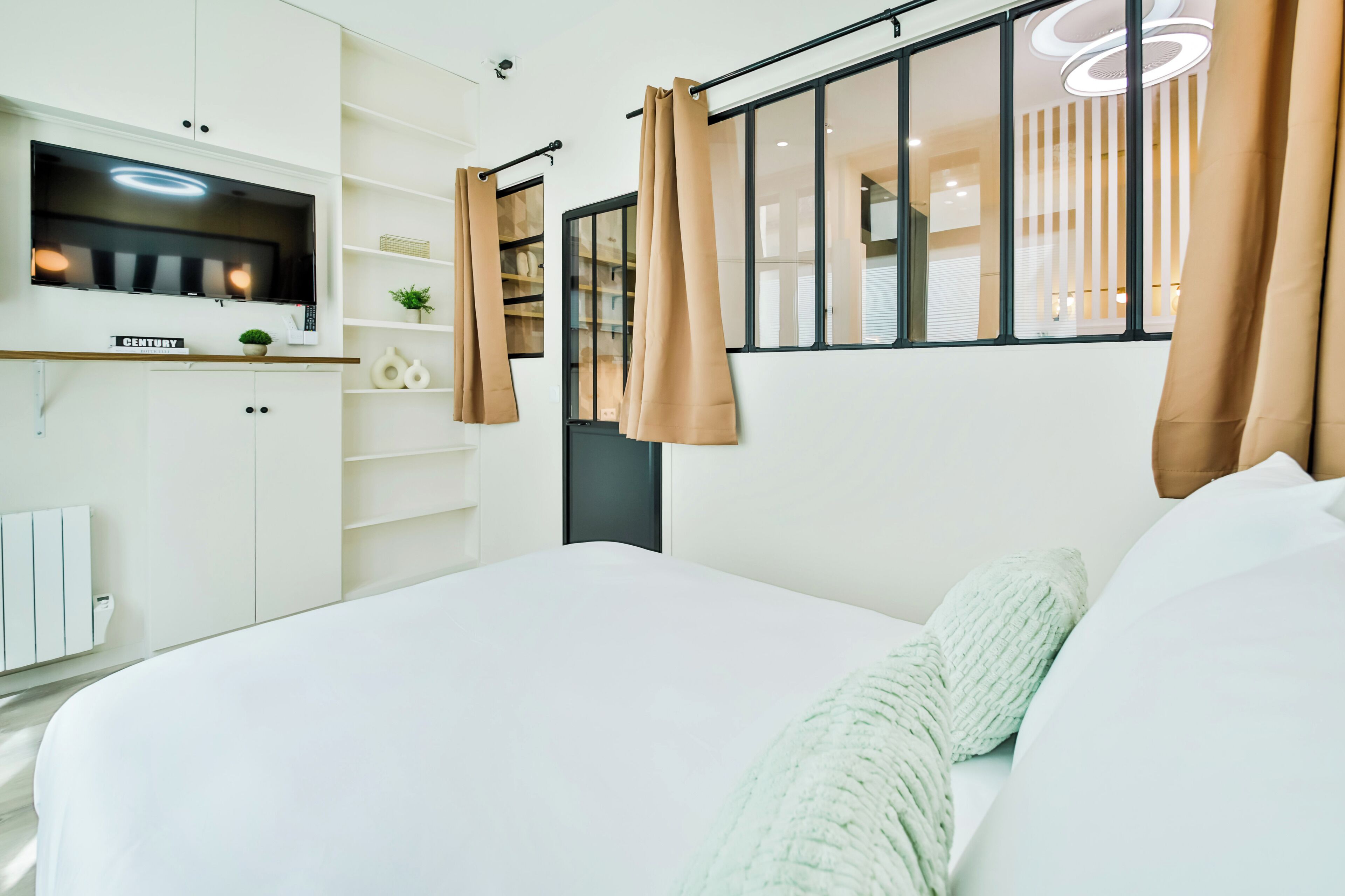 A modern bedroom with a large bed, a TV mounted on the wall, and a window with curtains.