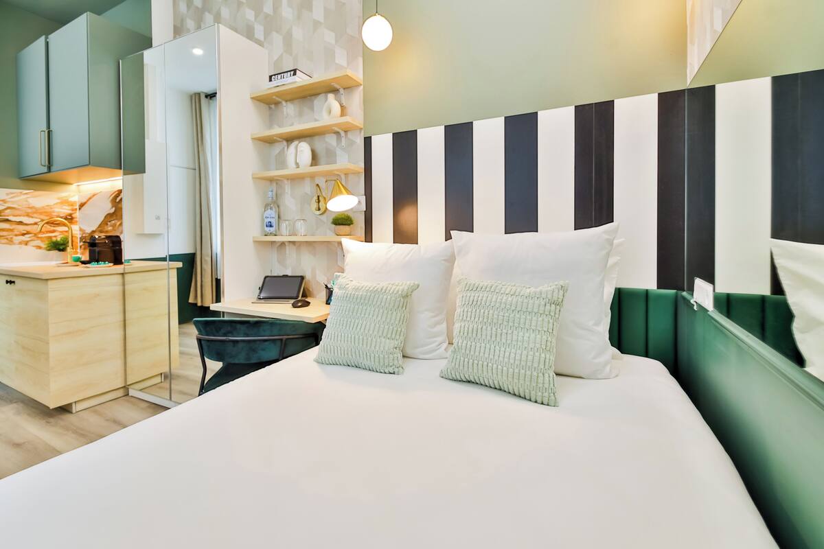 A modern hotel room with a large bed, a desk with a laptop, and a green accent wall.