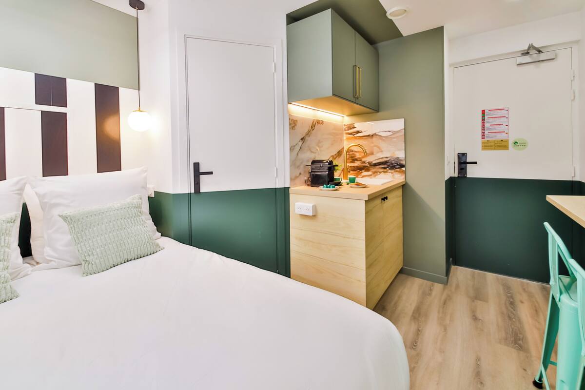 A modern hotel room with a bed, a small kitchenette, and a door.