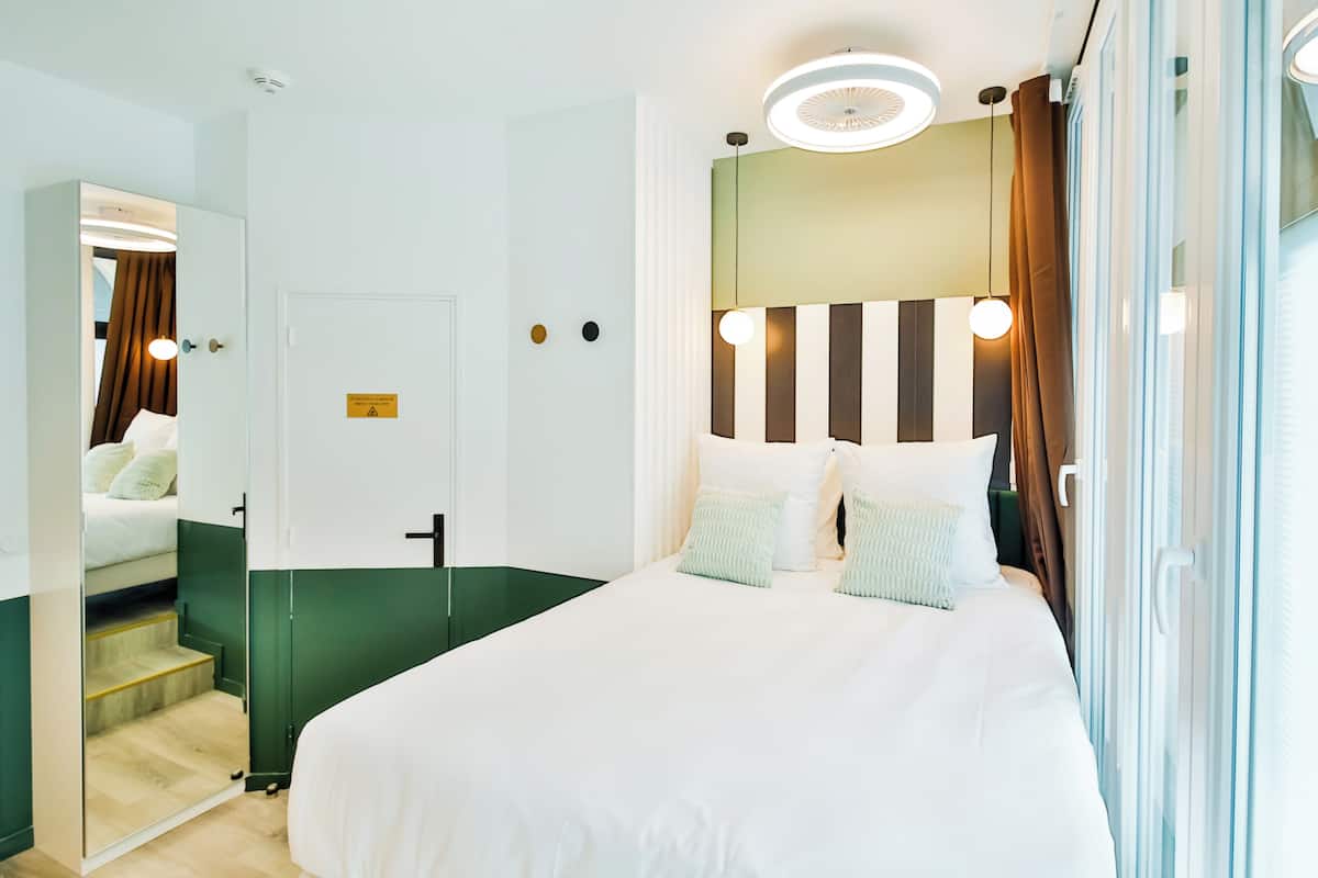 A modern hotel room with a large bed, a mirror, a green accent wall, and a view of the outdoors.