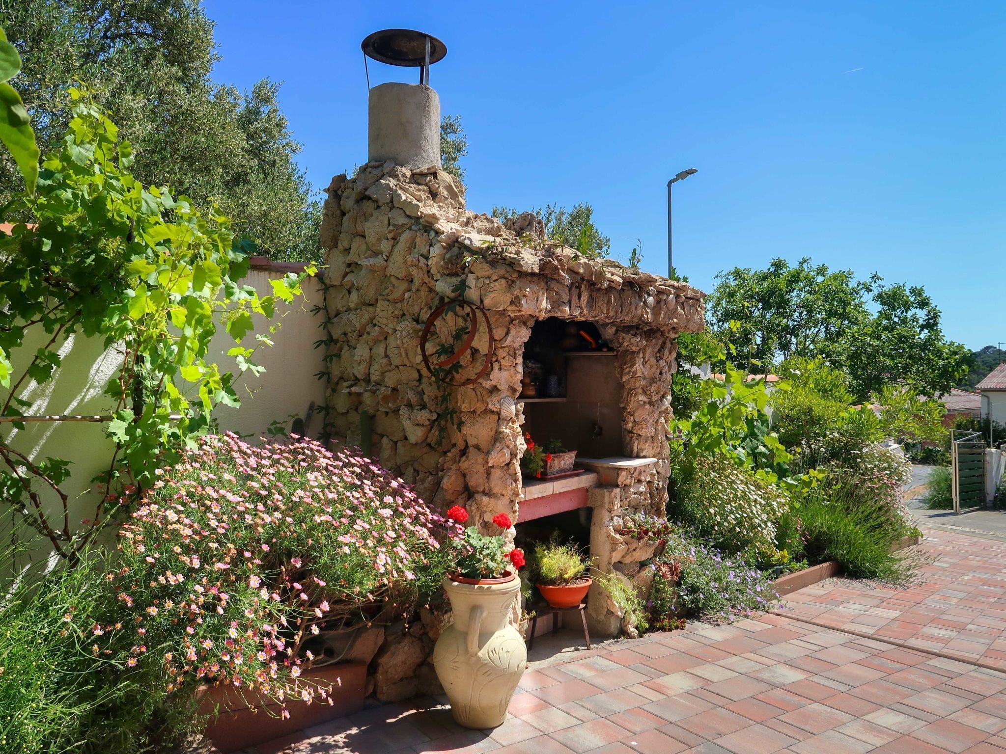 A stone-built outdoor kitchen with a chimney, surrounded by potted plants and flowers.
