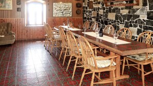 A dining area with a long wooden table set for six, surrounded by wooden chairs with cushioned seats. A stone wall with a wooden shelf above the table displays various items. - Vacation home Delfar by Interhome (Lošinj / Veli Lošinj)