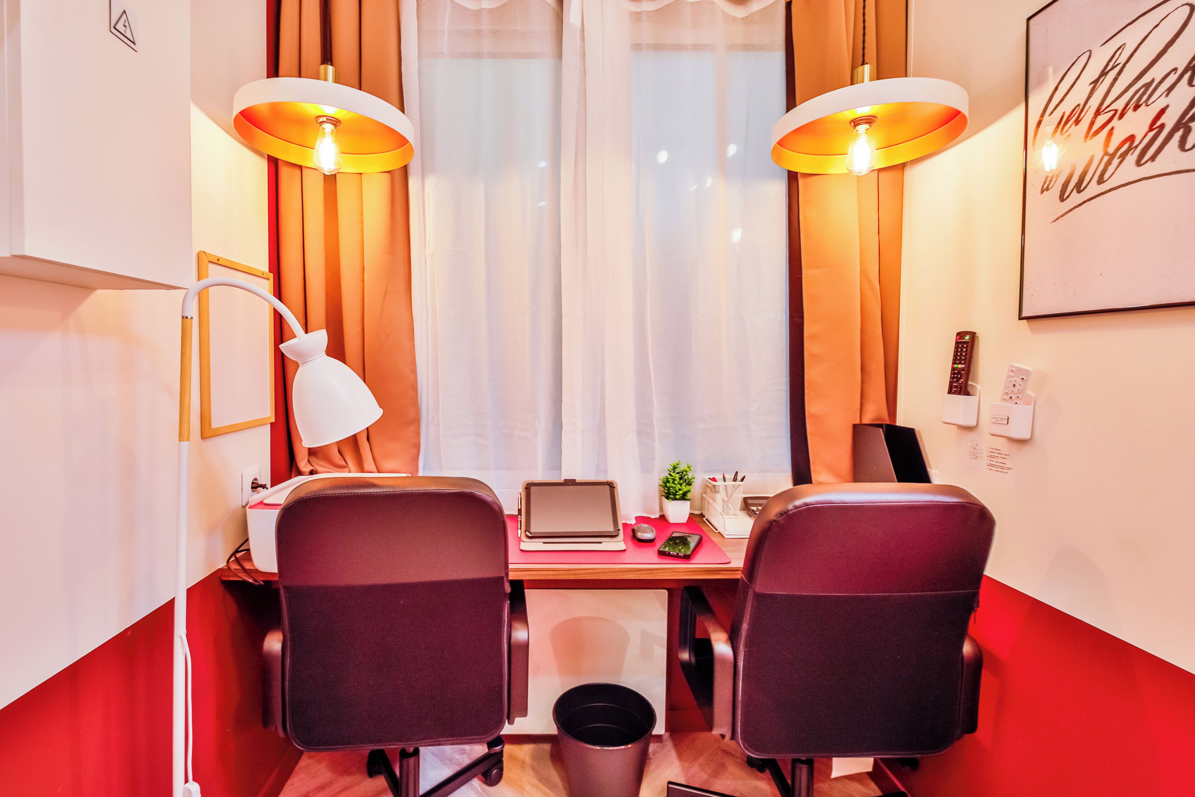 A hotel room with a desk, two chairs, a laptop, a plant, and a wall-mounted light fixture.