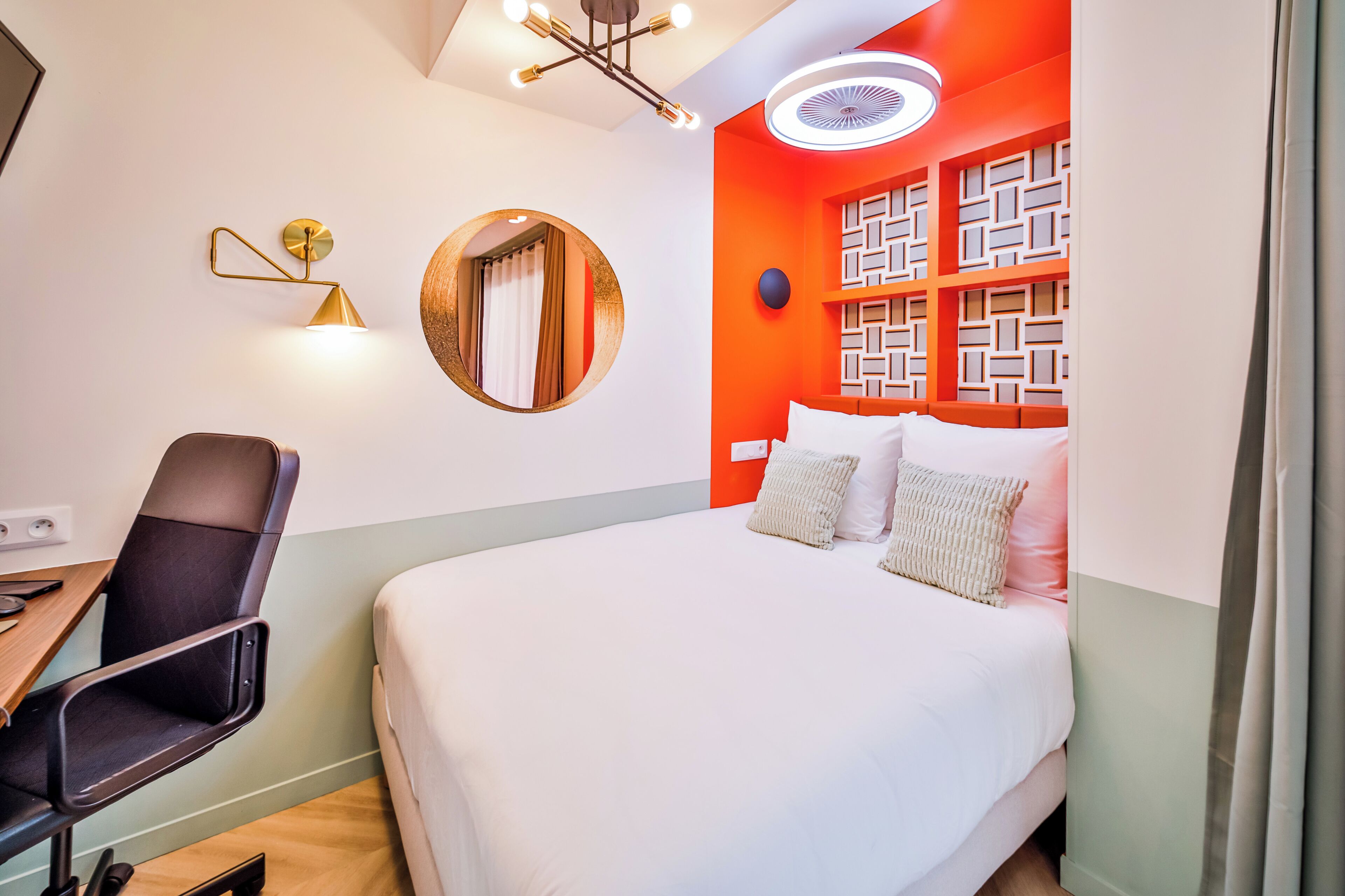 Una habitación de hotel moderna con una cama grande, un escritorio con una silla, un espejo redondo y un diseño de pared geométrico único.