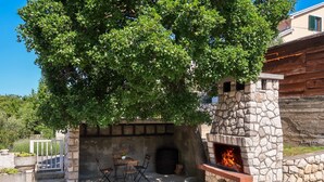A stone-built outdoor kitchen with a fire feature, surrounded by greenery and a seating area with chairs. - Apartment Monja by Interhome (Novi Vinodolski)
