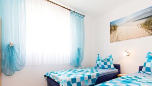 Ein Zimmer mit zwei Betten, Holzboden, einem Strandbild und einem Fenster mit durchsichtigen Vorhängen.