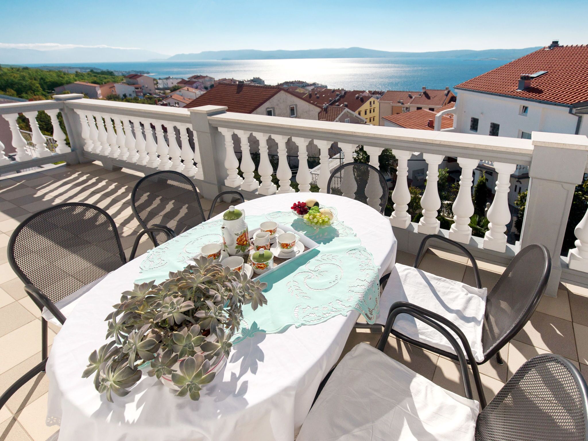 A terrace with a table set for tea, surrounded by chairs, overlooking a coastal town.