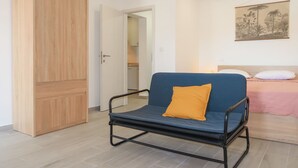 Una habitación con un armario de madera, un sofá azul con un cojín naranja, una cama con ropa de cama rosa y un cuadro colgado en la pared.