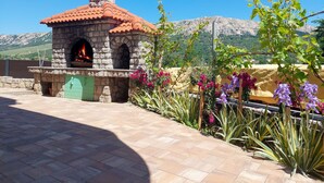 An outdoor stone oven with a terracotta roof, surrounded by flowering plants and a view of mountains. - Apartment Marija by Interhome (Krk/Baška)