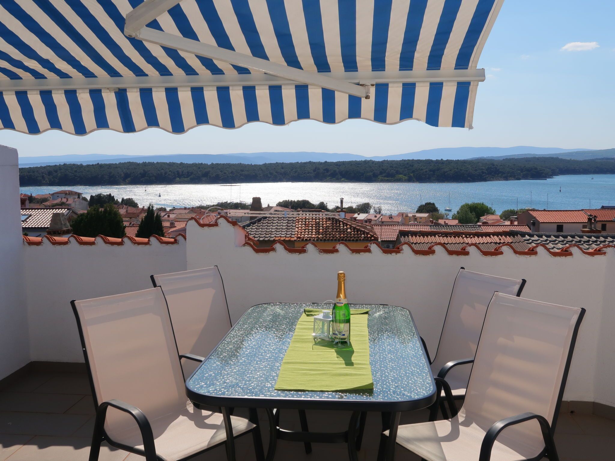 Mesa com cadeiras sob um toldo listrado, com vista para um lago e uma cidade.