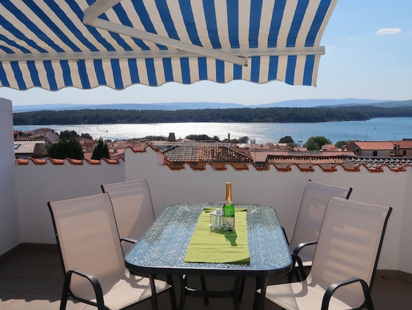 A table with chairs under a striped awning, overlooking a lake and town.