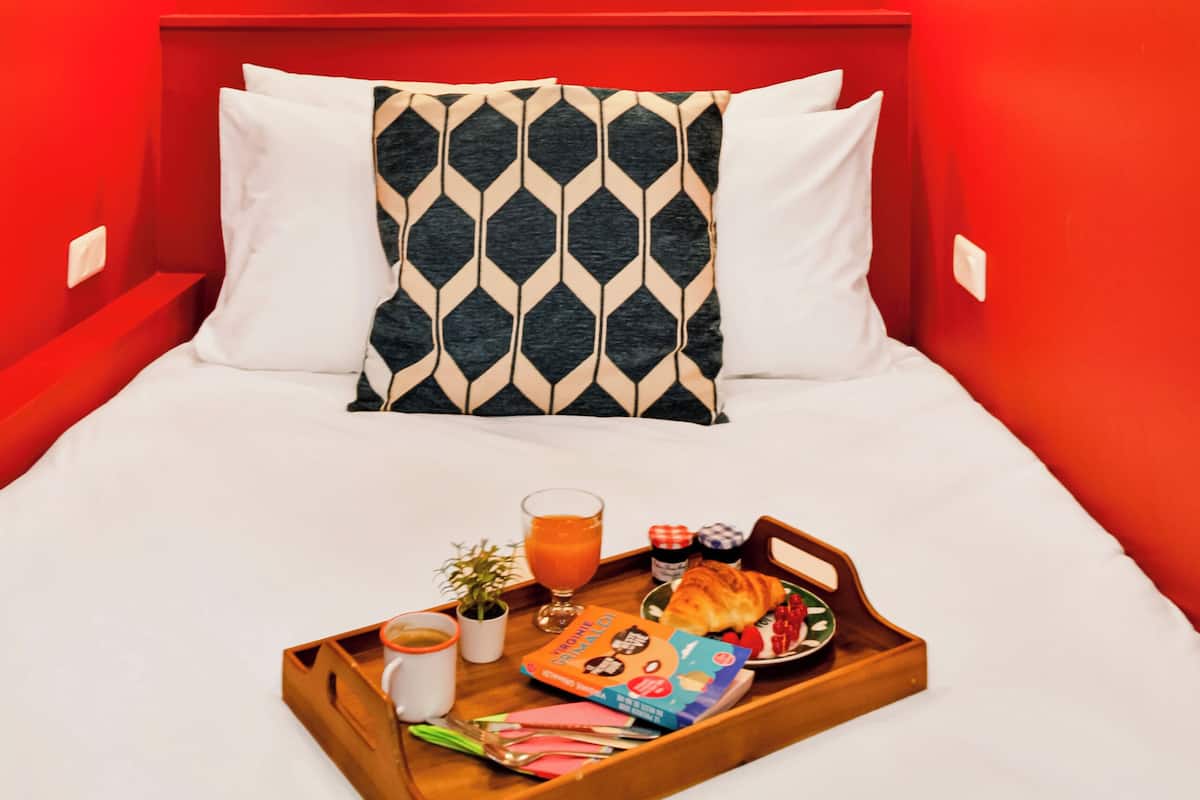 Habitación de hotel con pared roja, cama con ropa blanca, bandeja con alimentos para el desayuno y dos luces fijadas a la pared.