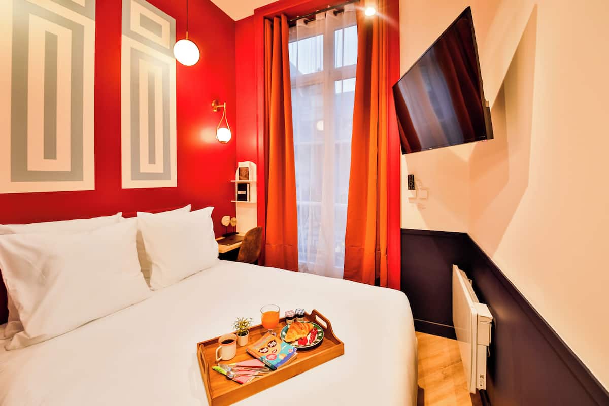 Habitación de hotel con una cama grande, una mesita de noche con lámpara, una bandeja con cosas para el desayuno y una ventana con cortinas.