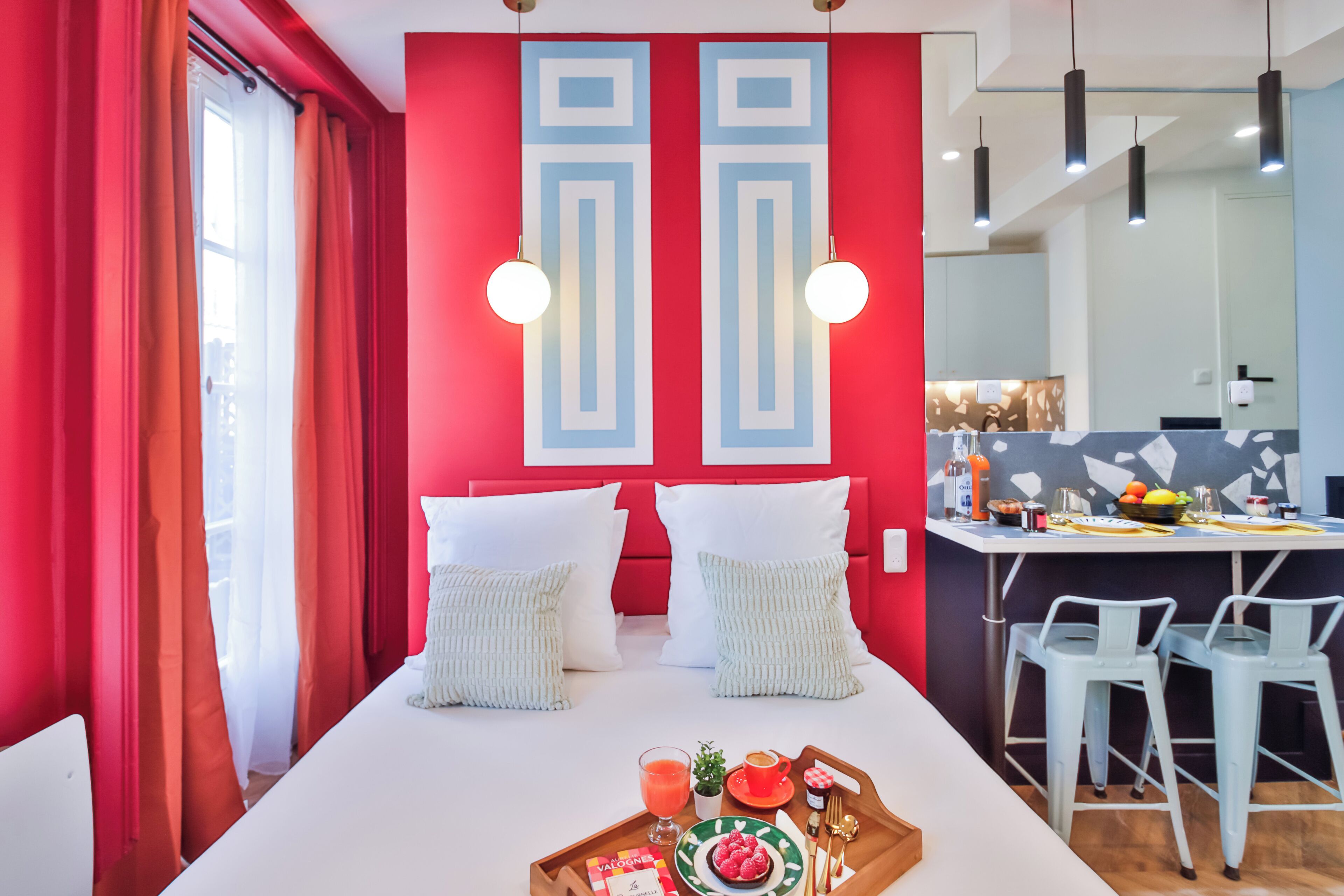 A modern hotel room with a red accent wall, a white bed with two pillows, a breakfast tray with food and drinks, and a kitchen area with bar stools and a counter.
