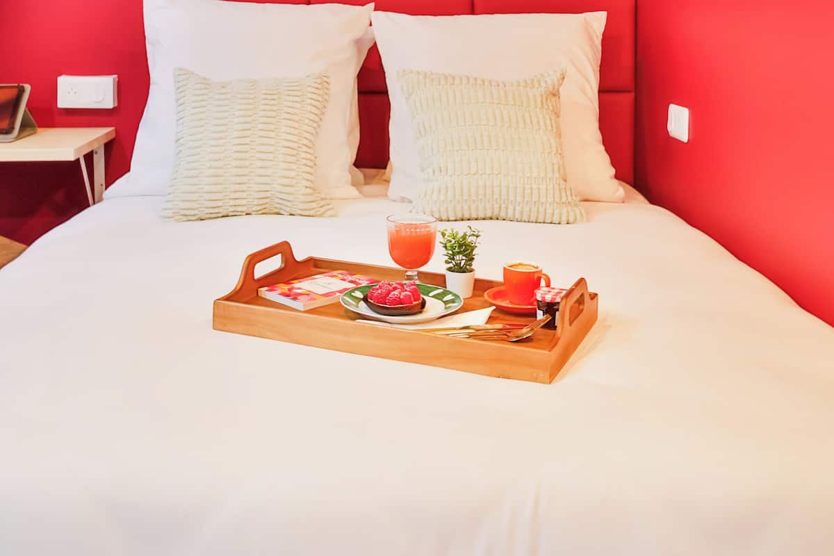 Una cama bien hecha con una bandeja de desayuno, una mesita de noche con lámpara y una pared roja con arte geométrico.