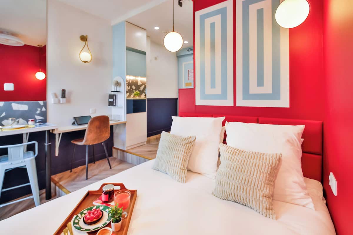 Habitación de hotel moderna con una pared de acento roja, cama con ropa de cama blanca, escritorio con silla y una bandeja con comida y bebida.