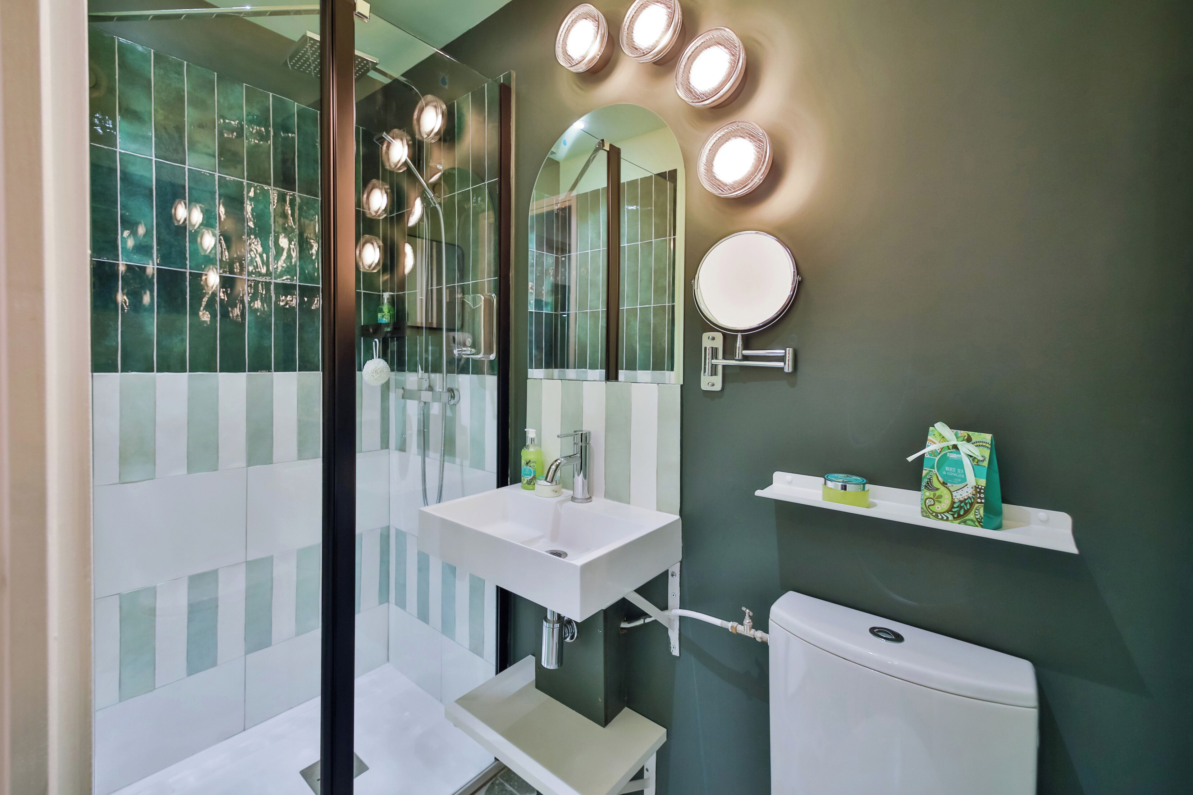 A modern bathroom with a white sink, a mirror, and a toilet. There are lights above the mirror and a shelf with toiletries.