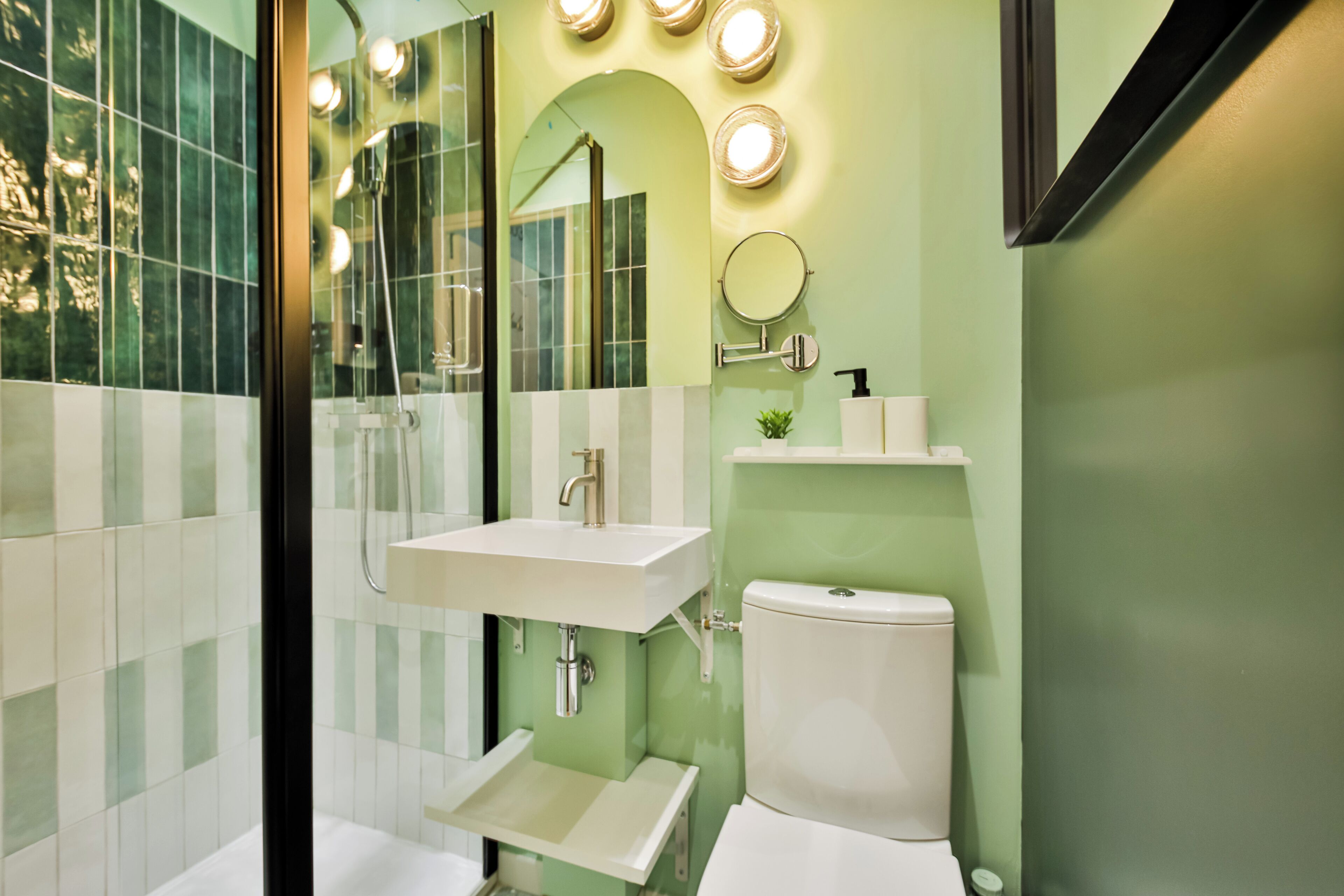 A bathroom with a glass shower enclosure, a white sink, a toilet, and a mirror with lights above it.