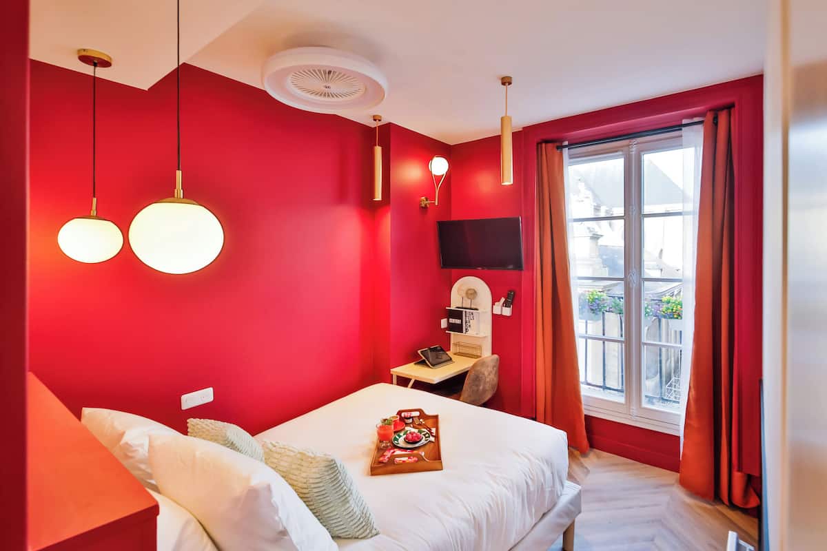 Habitación de hotel con pared roja, cama con ropa de cama blanca, escritorio con computadora, televisor y ventana con cortinas.