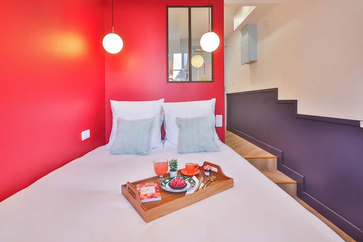 Habitación de hotel con una pared roja, una cama con almohadas blancas y azul claro, una bandeja con artículos de desayuno y una escalera.