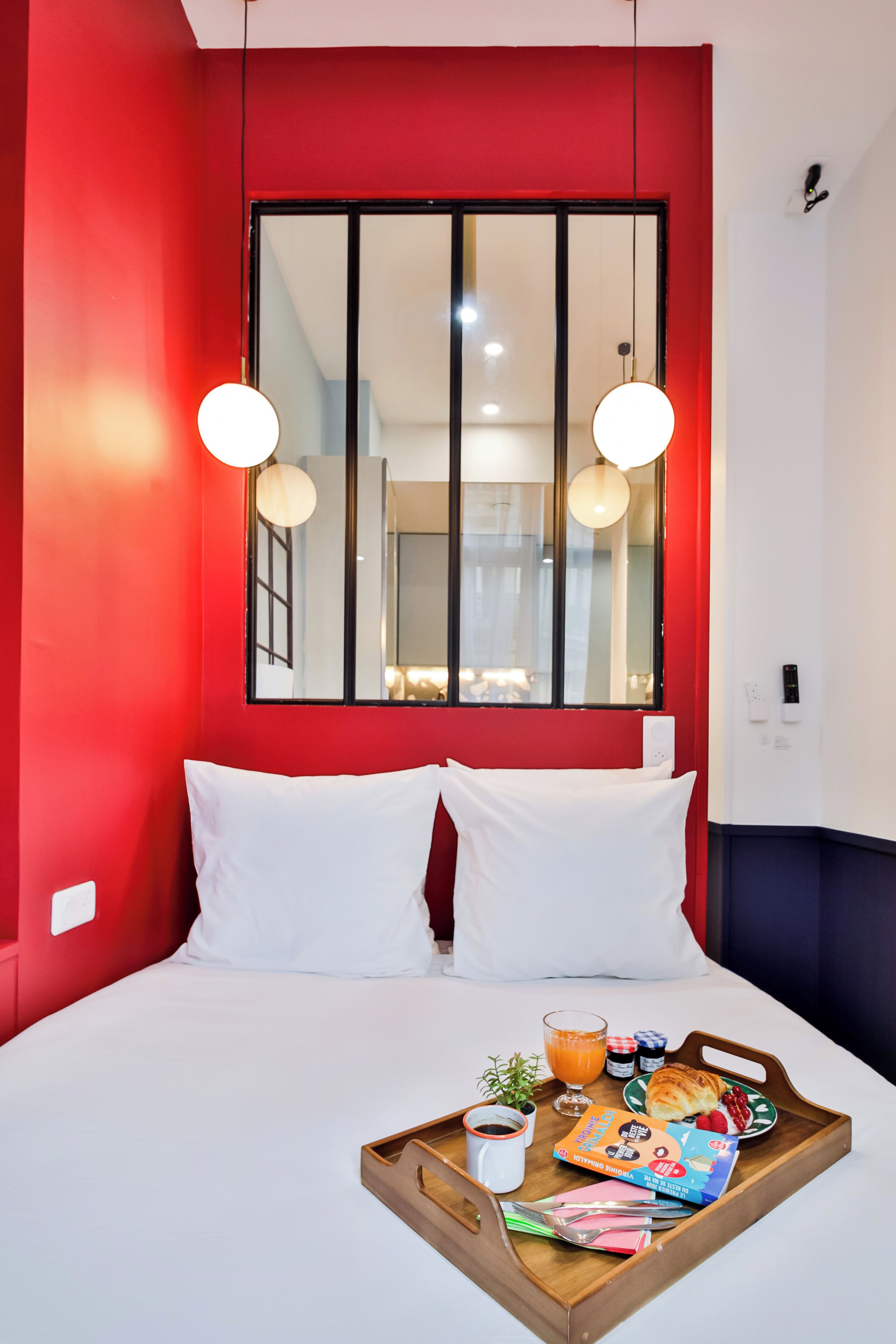 Une chambre d’hôtel avec un mur d’accent rouge, un grand miroir, du linge de lit blanc et un plateau de petit-déjeuner contenant divers articles.