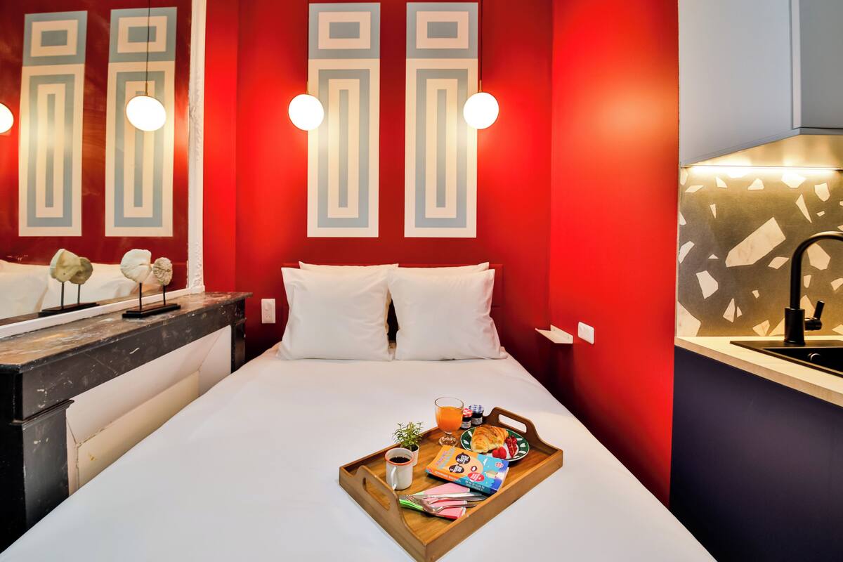 Una habitación de hotel con una cama, una bandeja con alimentos para el desayuno, una mesita de noche y una pared roja con diseños geométricos.