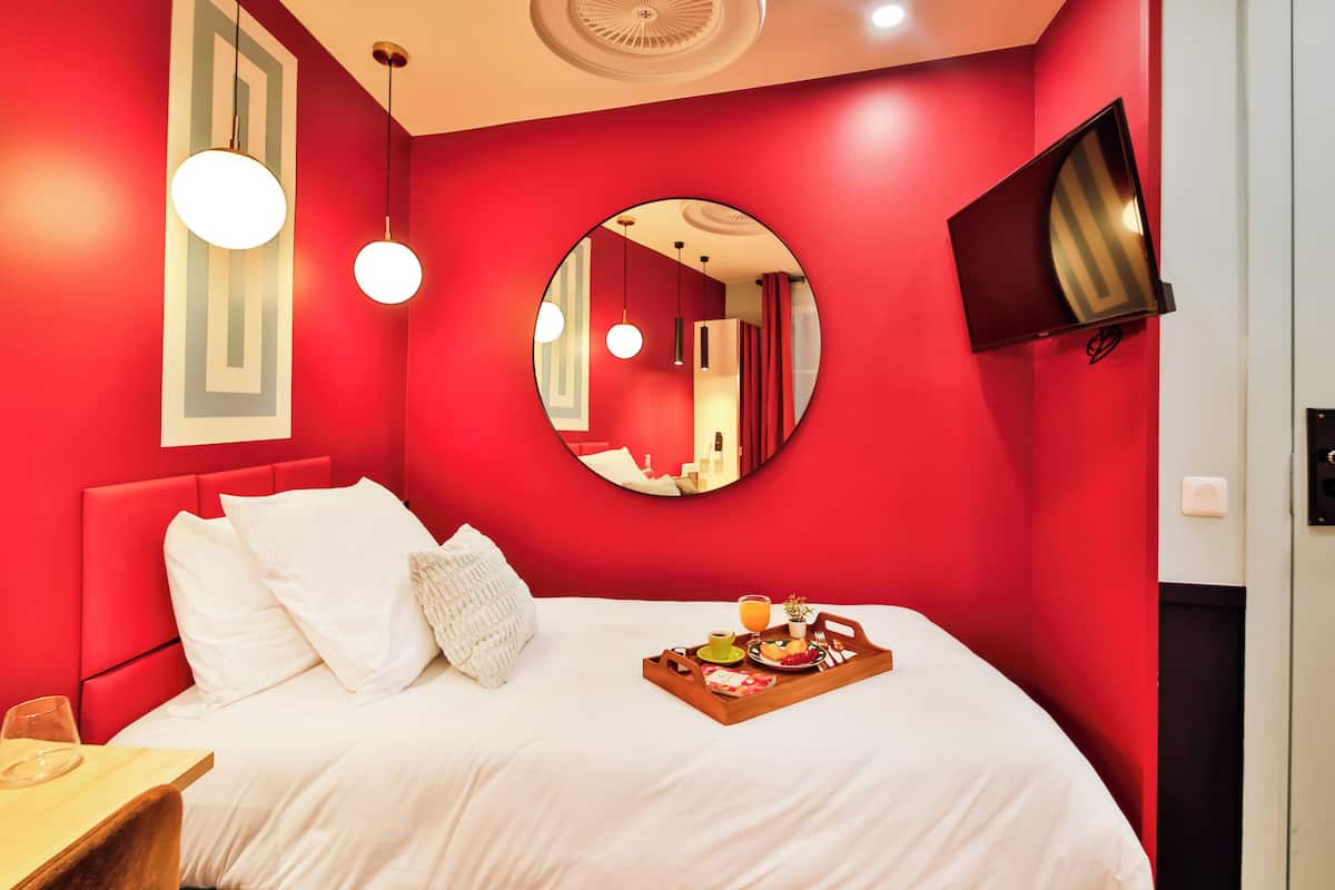 Un dormitorio con paredes rojas, una cama, ropa de cama blanca, un espejo redondo, un televisor de pantalla plana y una bandeja para el desayuno sobre la cama.
