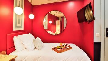 A red-walled bedroom with a bed, white bedding, a round mirror, a flat-screen TV, and a breakfast tray on the bed.