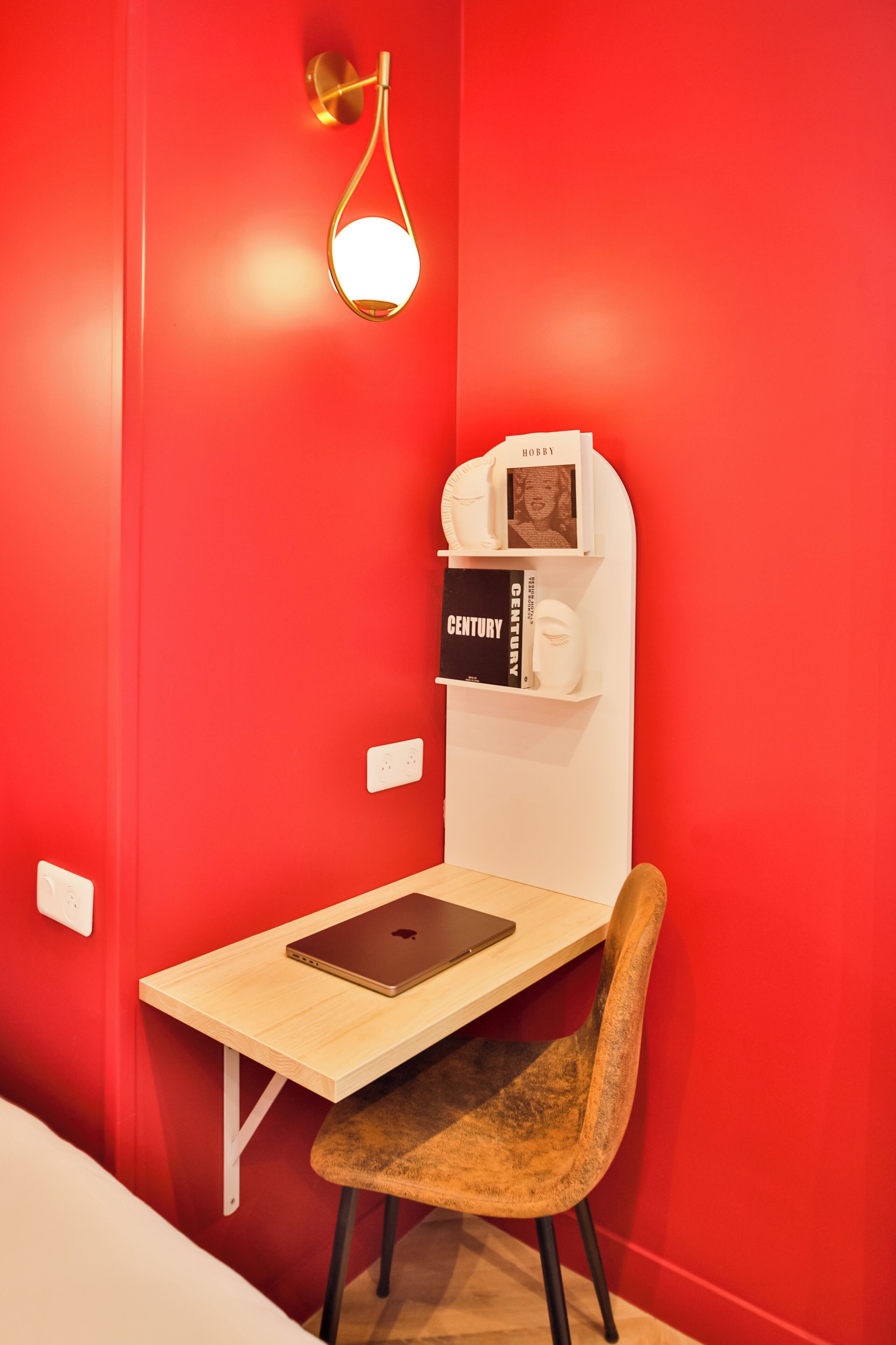 Una habitación con pared roja, un escritorio de madera con una computadora portátil, una silla, un estante en la pared con libros y un marco de fotos, y una lámpara de pared.
