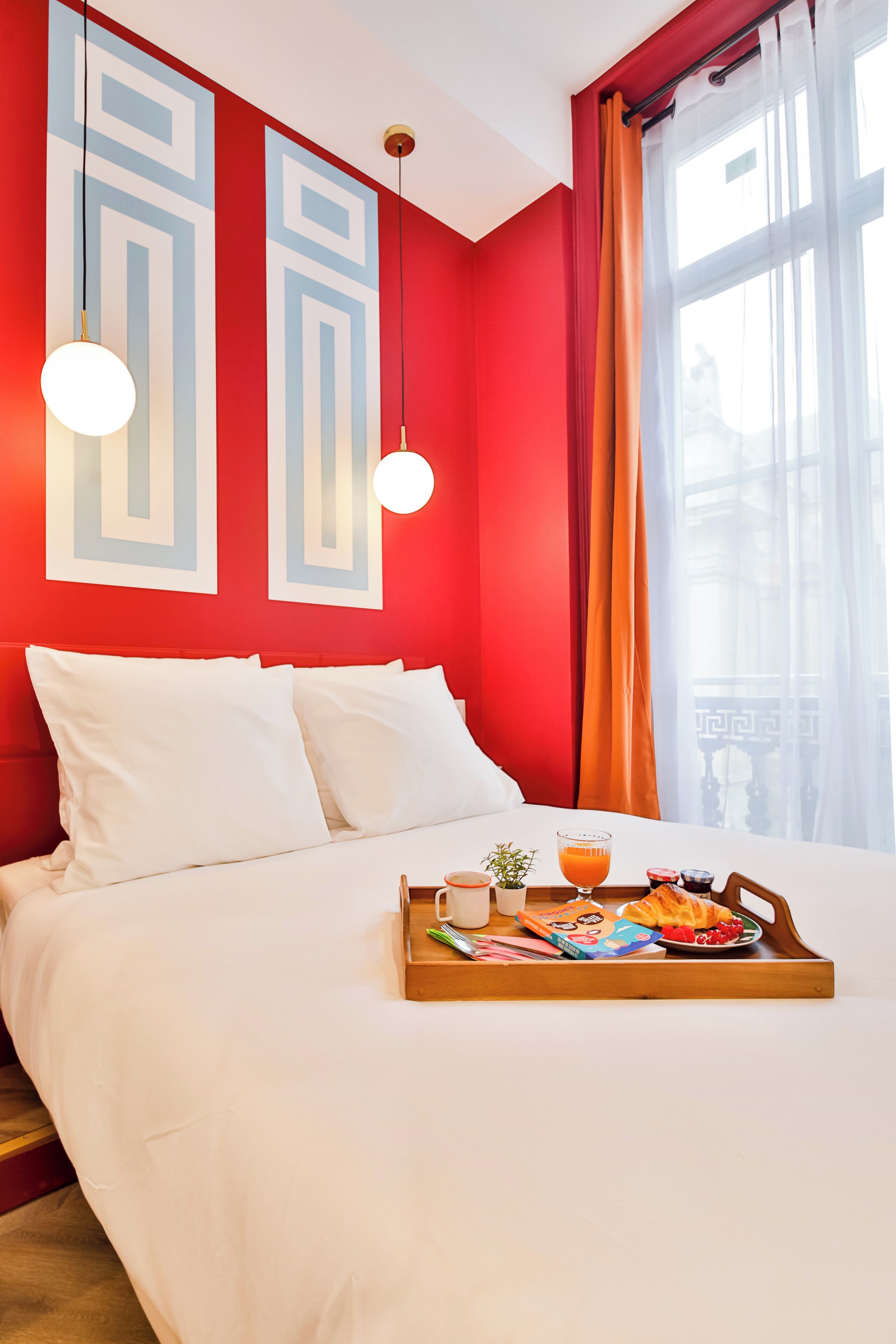 A hotel room with a red and white color scheme, a bed with white bedding, a tray with breakfast items, and a window with curtains.