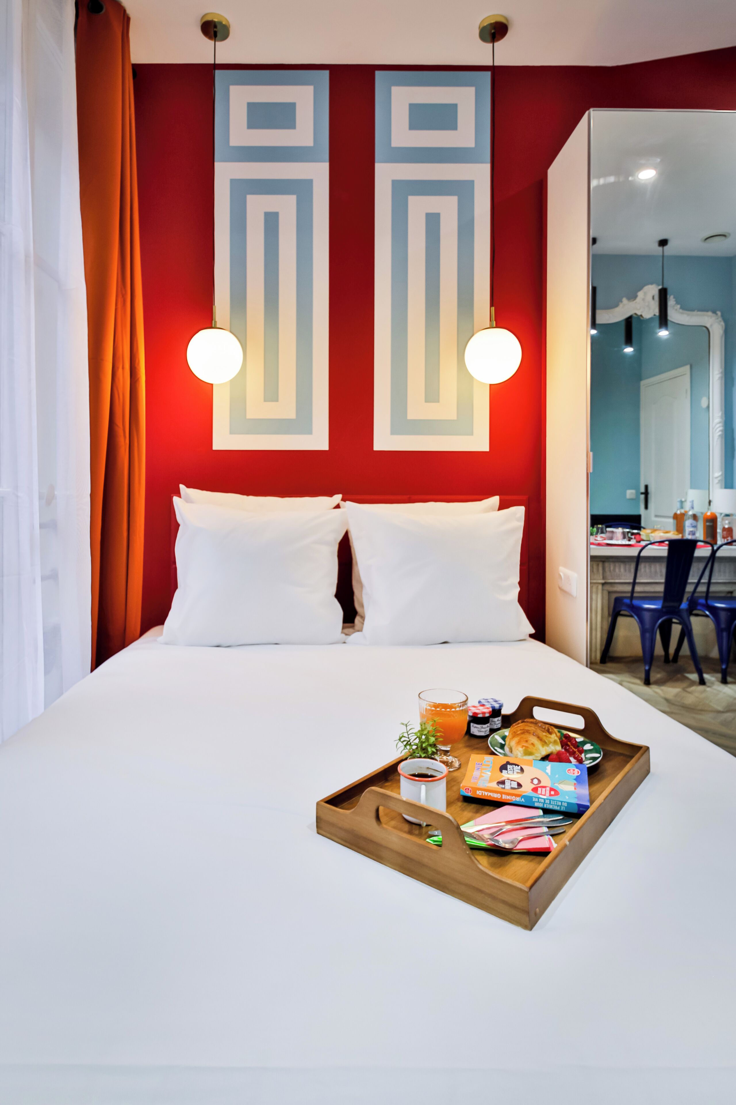 A hotel room with a bed, a tray with breakfast items, and a red wall with geometric patterns.