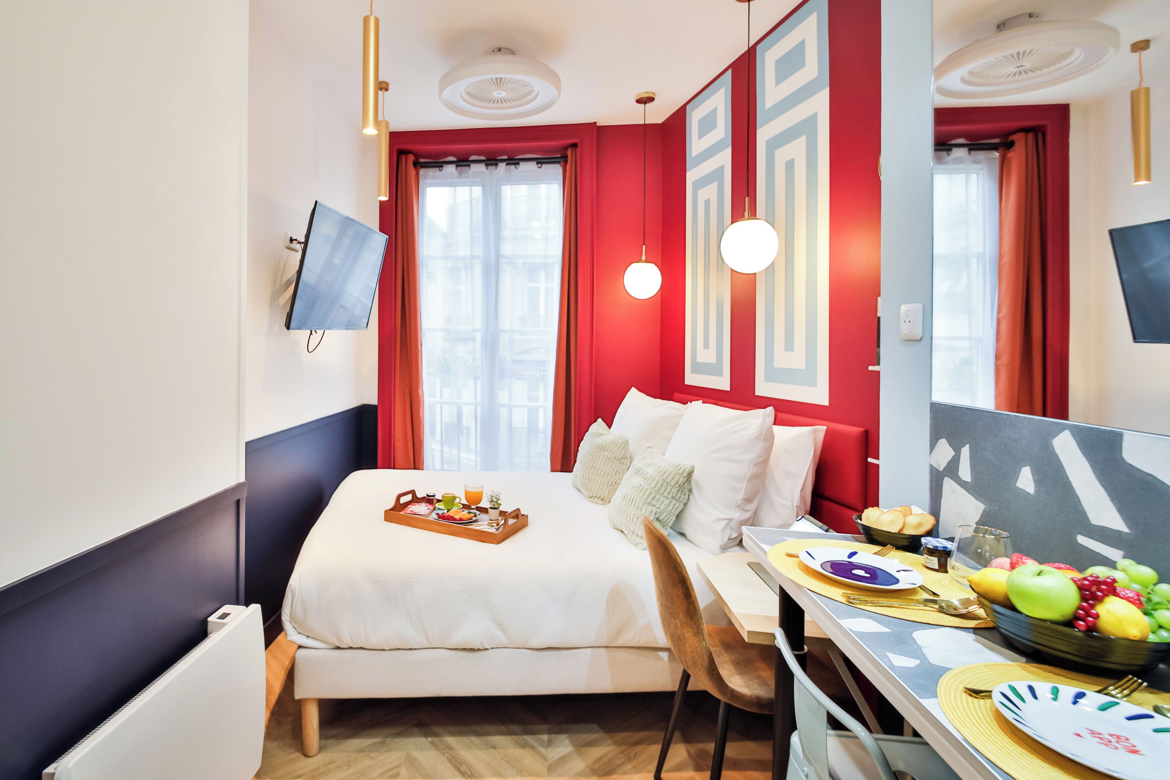 A hotel room with a bed, a desk with breakfast, a TV, and a window with curtains.