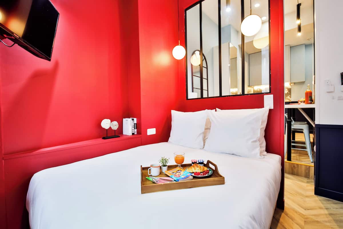 Habitación de hotel con pared de acento roja, una cama grande con ropa blanca, una bandeja con artículos de desayuno y un espejo sobre la cama.
