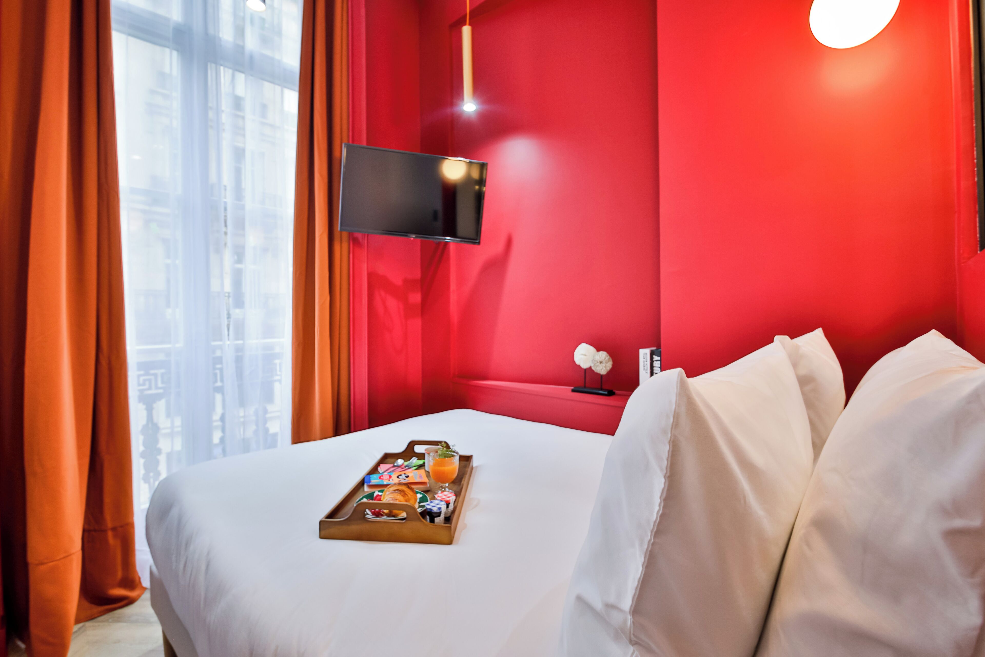 A hotel room with a red accent wall, a flat-screen TV mounted above the bed, and a breakfast tray on the bed.
