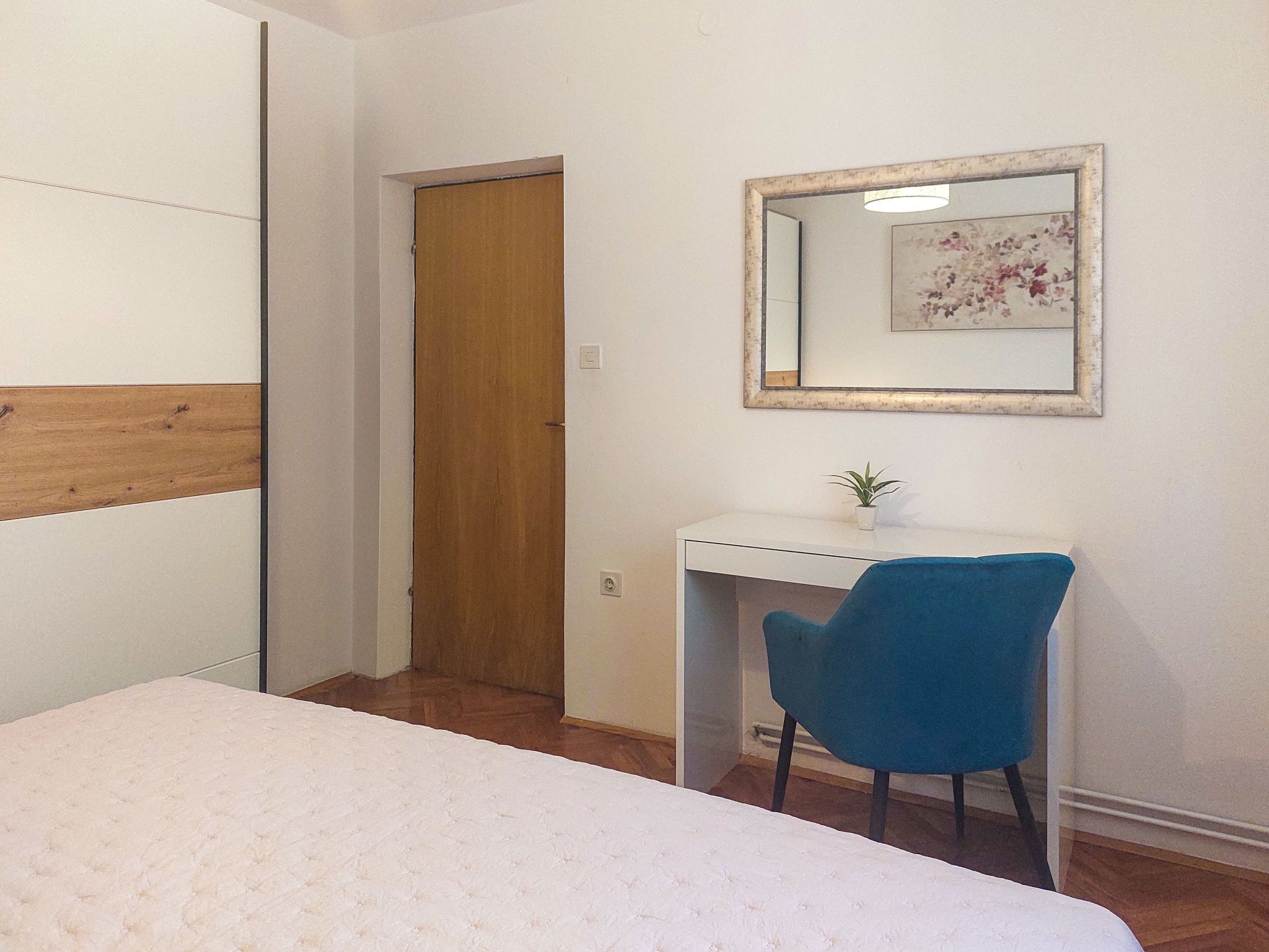 A bedroom with a bed, a desk, a chair, a mirror, and a door.