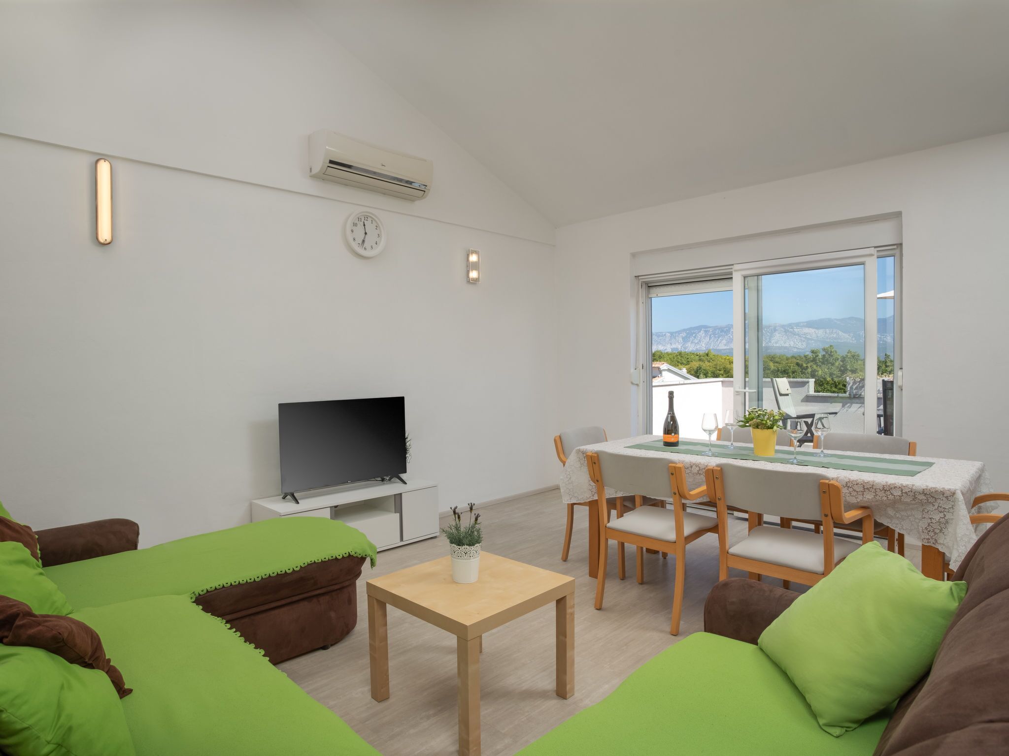 Un soggiorno moderno con una TV a schermo piatto, divani verdi, un tavolino in legno e una zona pranzo con tavolo e sedie.